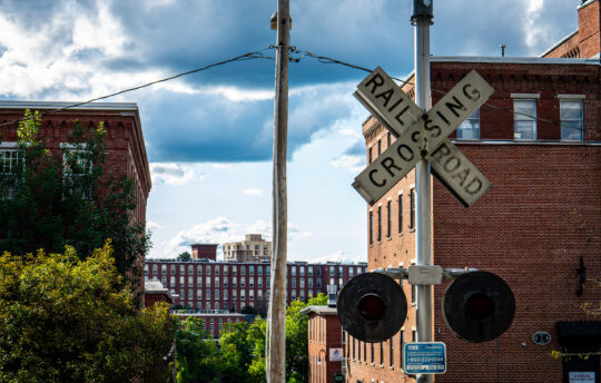 Railroad Crossing sign with two mill buildings behind it was a big open cloudy sky
