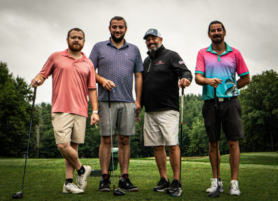 Four men on a golf course holding golf clubs with different colored shirts
