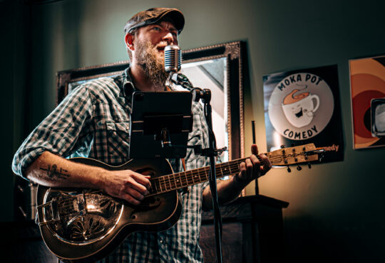 Man with a beard playing Acoustic-Electric Guitar. Singing into an old timey microphone with an ipad in front of him. He is standing infront of a piano