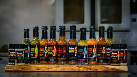 Hot sauces and salts on a cutting board on a metal cutting board in a professional kitchen with an oven in the background